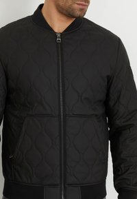 Chaqueta bomber acolchada negra con cierre de cremallera, cuello y puños de canalé, y bolsillos laterales; presenta un patrón de costura en ondas.