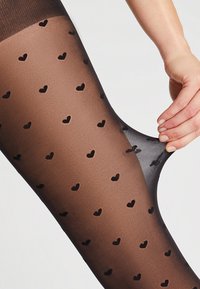 Collant marroni trasparenti con un motivo di piccoli cuori neri. Il materiale sembra elastico, liscio e semi-trasparente. Mano che afferra il tessuto.