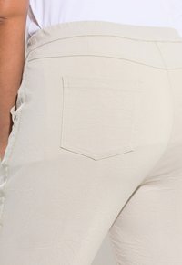 Pantaloni elasticizzati beige chiaro con una finitura texturizzata, dotati di una tasca sul retro e dettagli sulle cuciture laterali. Tasche elastiche in vita per comfort.