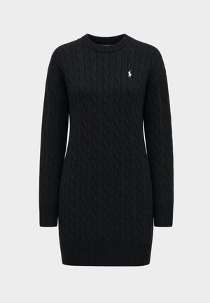 Femme aux cheveux bouclés et foncés portant une robe noire en maille à manches longues et des baskets blanches, debout devant un fond clair uni.