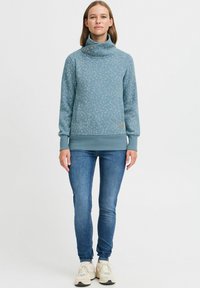 Sudadera azul claro con cuello alto, con un estampado floral, puños acanalados y un pequeño detalle de logo, llevada con jeans azules.