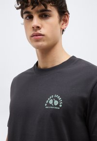 Νέος άνδρας που φοράει σκουρόχρωμο γκρι μπλουζάκι με ανοιχτό πράσινο γράμμα "MOPD SPORTS" και γραφικό ρακέτας τέννις στο στήθος, με απλό φόντο.