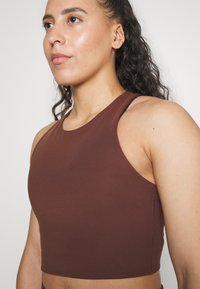 Smeđi, priljubljeni sportski crop top s visokim izrezom i širokim trakama, napravljen od rastezljivog materijala. Glatka tekstura s urednim šavovima.