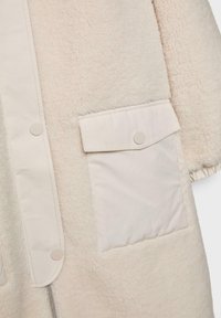 Kremowy polarowy płaszcz z gładką beżową kieszenią na patkę, zatrzaskami oraz miękką teksturą. Posiada zamek błyskawiczny i marszczone zakończenia rękawów.