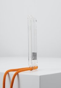 Clear mobile phone case with orange strap. Features a transparent design and logo "URBAN CLASSICS" on the back. Textured strap for grip.