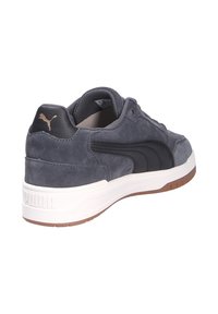Sneaker in suede grigio scuro con dettaglio in pelle nera, suola intermedia bianca e suola esterna marrone. Presenta un logo dorato sul tallone. Design classico.