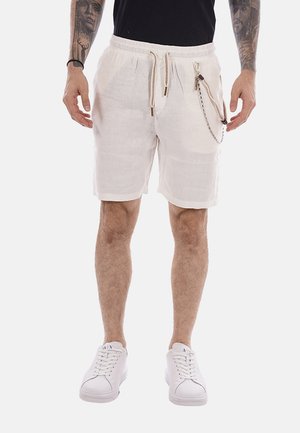 Uomo che indossa pantaloncini bianchi con coulisse, zip e dettaglio a catena, maglietta nera, scarpe da ginnastica bianche e tatuaggi su entrambi gli avambracci.