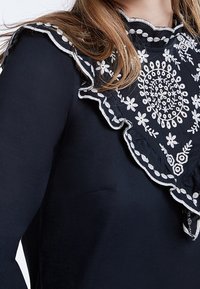 Sort beklædning med en dekorativ hvid broderet krave med blomster mønstre og bølgede kanter, der fremhæver detaljerigdom og tekstur.