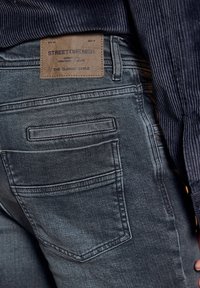 Zbliżenie na ciemnoszare dżinsy z tylną kieszenią, na której znajduje się beżowa skórzana metka z napisem "Street One Men", oraz część prążkowanego granatowego swetra.