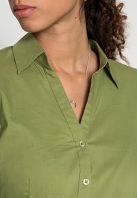 Camicia verde con bottoni, dotata di un colletto strutturato, caratterizzata da una texture liscia e piccoli bottoni. È visibile una collana a forma di falce di luna in argento.