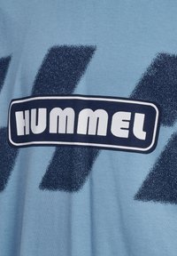 Lyseblå bomulds t-shirt med et stort navy og hvidt "HUMMEL" logo, prydet med teksturerede abstrakte mønstre i mørkeblå.