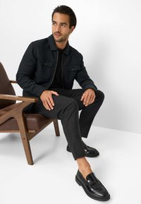 Zwarte ritsjack met voorzakken, gecombineerd met donkergrijze pantalon en zwarte instap-schoenen. Zittend op een houten stoel tegen een witte achtergrond.