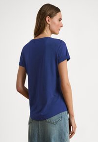 Γυναίκα που φοράει μπλε κοντομάνικο πουκάμισο και ανοιχτόχρωμη τζιν φούστα, απεικονιζόμενη από πίσω με φόντο ένα απλό λευκό τοίχο.