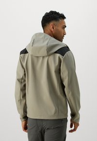 Vaaleanbeige softshell-takki hupparilla, jossa on mustat olkatoppaukset ja teksturoitu pinta, päälle puettuna harmaiden housujen kanssa.