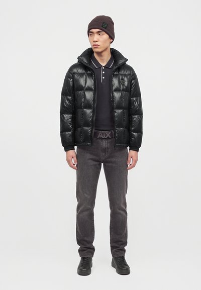 Jeune homme portant une doudoune noire, un polo noir, un jean foncé, des baskets noires, un bonnet en tricot marron, et une ceinture avec une boucle "A|X".