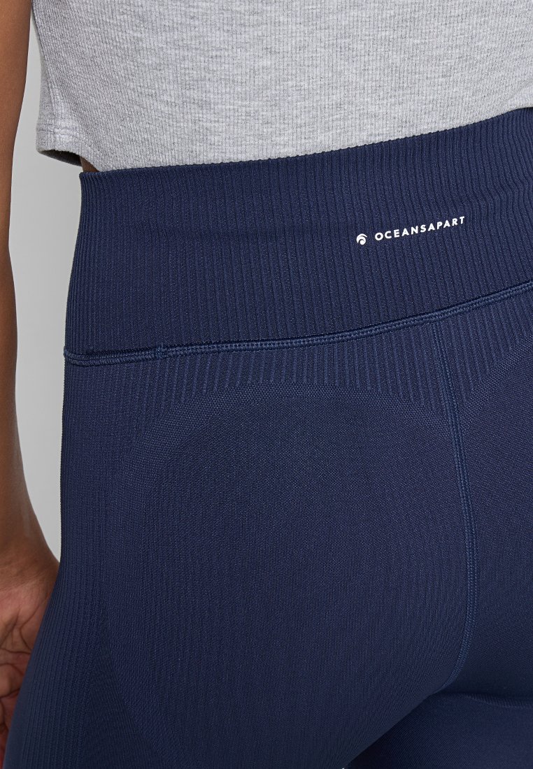 Nahaufnahme der Taille und Hüften einer Person, die marineblaue Rippleggings mit einem hohen Bund und einem kleinen weißen "OCEANSAPART"-Logo trägt.