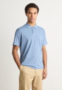 Cămașă polo de culoare bleu deschis, din bumbac, având un guler, trei nasturi și un detaliu subtil al logo-ului pe piept. Asortată cu pantaloni bej.