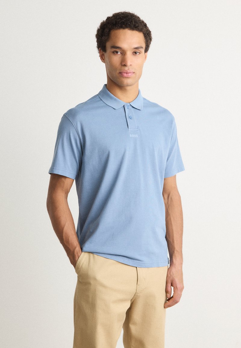 Cămașă polo de culoare bleu deschis, din bumbac, având un guler, trei nasturi și un detaliu subtil al logo-ului pe piept. Asortată cu pantaloni bej.