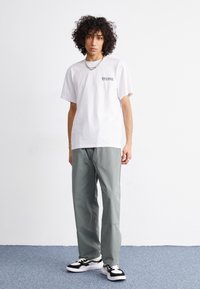 T-shirt bianca in cotone con una grafica nera sul petto, abbinata a pantaloni larghi di un verde chiaro. Le scarpe presentano accenti in bianco e nero.