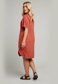 Rostfärgad klänning med korta ärmar, avslappnad passform och sidfickor. Buren med svarta sandaler med spänne. Slät textur.
