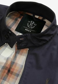 Veste marine avec col noir, doublure à carreaux beige, grise et orange, fermeture éclair sur le devant, et étiquette noire indiquant "1982 Authentic Design NEXT" avec un logo de cerf.