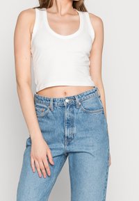 Top corto blanco sin mangas con cuello redondo combinado con unos jeans de mezclilla de tiro alto en azul claro que presentan un clásico diseño de cinco bolsillos.