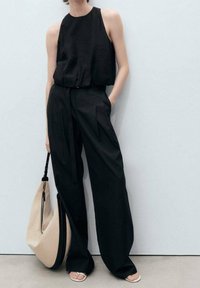 Haut noir sans manches en tissu texturé, associé à un pantalon large noir. Accessoirisé d'un grand sac beige et noir souple.