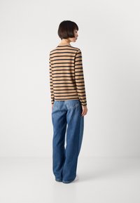 Donna con capelli corti che indossa una camicia a maniche lunghe a strisce beige e nere e jeans blu a gamba larga, in piedi di spalle su uno sfondo bianco.