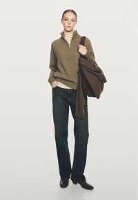 Groene kabelgebreide sweater met halve rits, crèmekleurige coltrui eronder, donkerblauwe jeans en een bruine tas. Model staat met handen in de zakken.