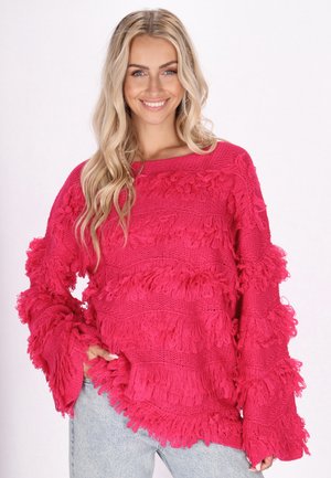 Fuchsia oversized trui met gestructureerde franjes, lange mouwen en een brede halslijn. Gecombineerd met lichtgekleurde spijkerbroek.
