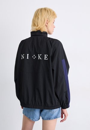 Zwarte windbreakerjas met het "NIKE"-logo op de rug in het wit. Bevat paarse accenten op de mouwen en een elastische onderkant en manchetten.