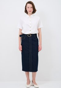 Camisa blanca de manga corta con botones dorados, combinada con una falda lápiz de mezclilla oscura ajustada y zapatos de tacón blancos.