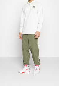 Polo blanco de manga larga con cremallera, logo de Nike en naranja, combinado con pantalones de chándal verde olivo con rayas blancas a los lados y zapatillas blancas con detalles en rojo.