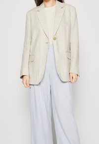 Beige lněný blejzr s jedním knoflíkem, klopovými límci a dvěma předními kapsami, nošený přes světlý top a světlé modré široké kalhoty.