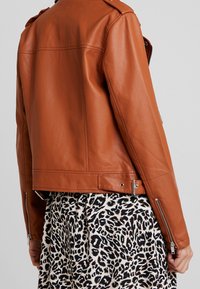 Giacca di pelle marrone con design aderente, colletto alla mandarina, tasche laterali con zip e hardware argentato. Indossata su una gonna con stampa leopardata bianca e nera.