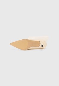 Tejpontú stiletto sarok krém színű patent anyagból, világos bézs talppal és fekete díszítéssel a sarok aljánál.