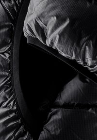 Den sorte pufferjakke har en glat inderside med vandret syninger. Kraven har en ribbet strikdetalje for øget isolering.