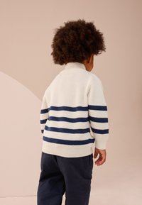 Maglione bianco a coste con righe orizzontali blu navy, collo alto e maniche lunghe. Abbinato a pantaloni scuri, visto da dietro.