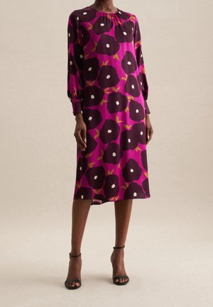Woman wearing a knee-length magenta dress with large dark purple floral patterns and black ankle-strap heels, standing against a plain background.