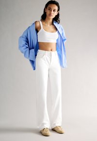 Jeans blancs à jambe large, débardeur blanc court et chemise oversize bleu clair ; présente une texture côtelée et des baskets beiges.