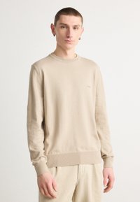 Beživi pulover, kroja z okroglim izrezom, rebrasti manšeti in spodnji rob, mehak material, minimalno označevanje, v kombinaciji s svetlimi kordovanskimi hlačami.