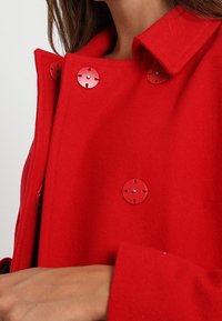 Abrigo de lana rojo con un cuello estructurado, adornado con grandes botones redondos rojos con un detalle en el centro plateado. Textura suave y silueta recta.