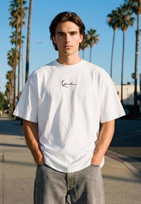 Mladić stoji na sunčanoj ulici s palmama, nosi bijelu preveliku majicu s natpisom "Kani" i sive traperice, ruke u džepovima.