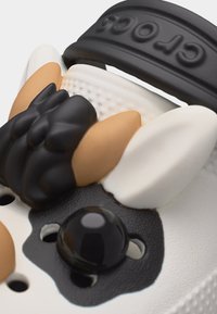 Κοντινό πλάνο ενός λευκού παπουτσιού Crocs με ένα μαύρο και καφέ γοητευτικό μοτίβο προσώπου μπουλντόγκ, προσαρτημένο κοντά στον λουρί της φτέρνας.