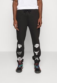 Svarta svettbyxor med elastisk midja, prydda med olika vita NBA-lagloggor tryckta längs benen. Kombineras med färgglada sportskor.