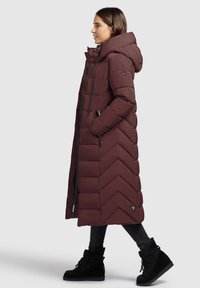 Lang bordeauxkleurige donsjas met een chevron-quilted patroon, een capuchon en zijzakken met rits, gecombineerd met zwarte enkellaarsjes.