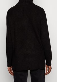 Maglione nero lavorato a maglia con colletto alto, vestibilità ampia e texture a costine. Presenta maniche lunghe e un orlo posteriore leggermente più lungo.