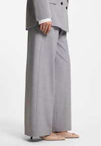 Ανοιχτό γκρι κοστούμι με παντελόνια φαρδιάς ποδός και ταιριαστό σακάκι διπλής σειράς κουμπιών. Λείο υφικό, μινιμαλιστικός σχεδιασμός και μυτερά μπεζ παπούτσια.