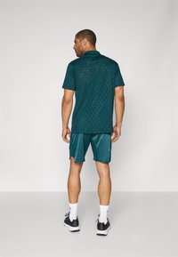Türkisfarbenes sportliches Poloshirt mit Diamantmuster, kombiniert mit passenden Shorts. Der Stoff hat eine glatte Textur; getragen mit schwarzen Sneakers und weißen Socken.