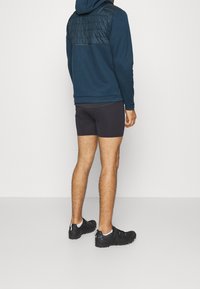 Hoodie bleu foncé avec un panneau matelassé sur la poitrine, associé à un short de sport noir. La personne porte des chaussures de sport noires avec des sangles sécurisées.